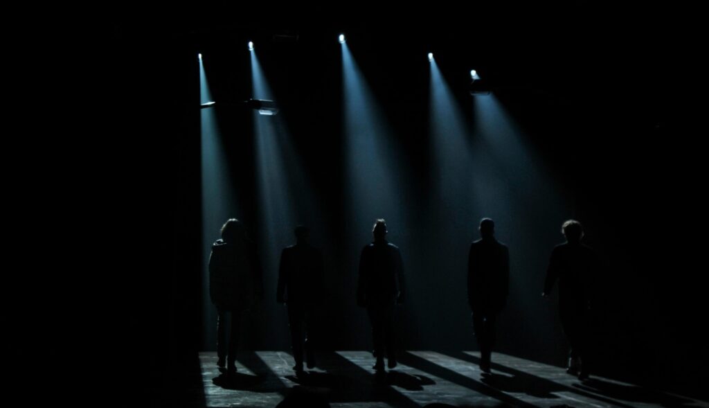 Silhouettes of people walking on stage beneath dramatic spotlights, creating a moody atmosphere.
