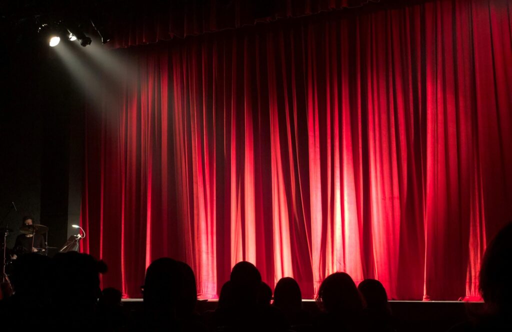 Dimly lit theater stage with red curtains and audience silhouettes under spotlights.